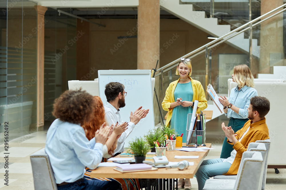 Young bright designers cheerfully applaud, sit at light coloured wooden table in cozy roomy office, horizontal shot, design agency concept Young bright designers cheerfully applaud, sit at light coloured wooden table in cozy roomy office, horizontal shot, design agency concept