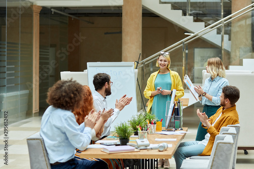 Fotografie Young bright designers cheerfully applaud, sit at light coloured wooden table in