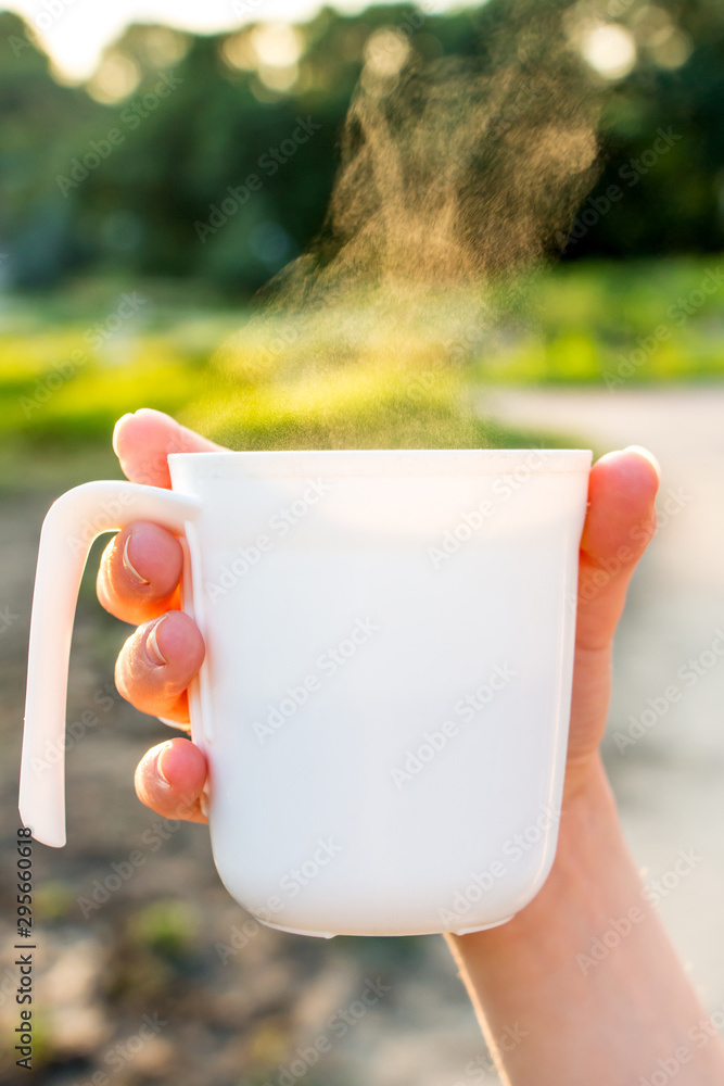Obraz premium a white plastic mug from which steam comes out in the hands of a young girl, lit by the rays of the morning sun against a background of nature