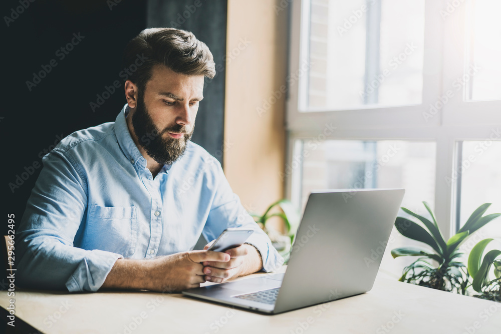 Young man sitting at window and working on laptop. Bearded man sitting at desktop with smartphone in his hands. Online education. Young man sitting at window and working on laptop. Bearded man sitting at desktop with smartphone in his hands. Online education.