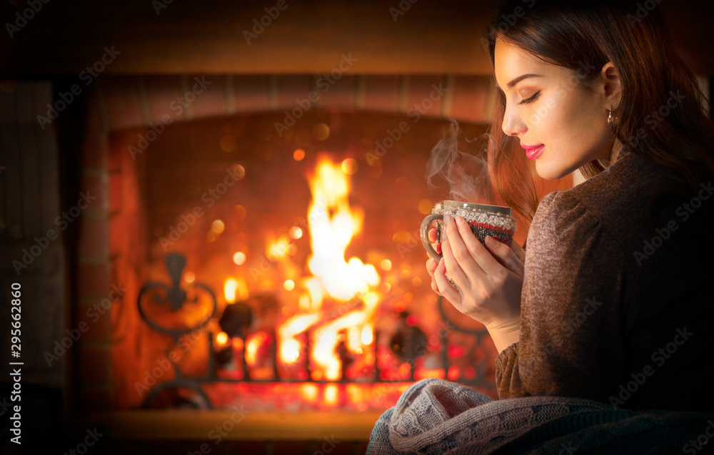 Beauty young Christmas woman sitting near fireplace in dark room at home and drinking hot beverage from a big cup. Beauty girl Enjoying tea, coffee or hot chocolate in a cozy room. Xmas scene - obrazy, fototapety, plakaty 