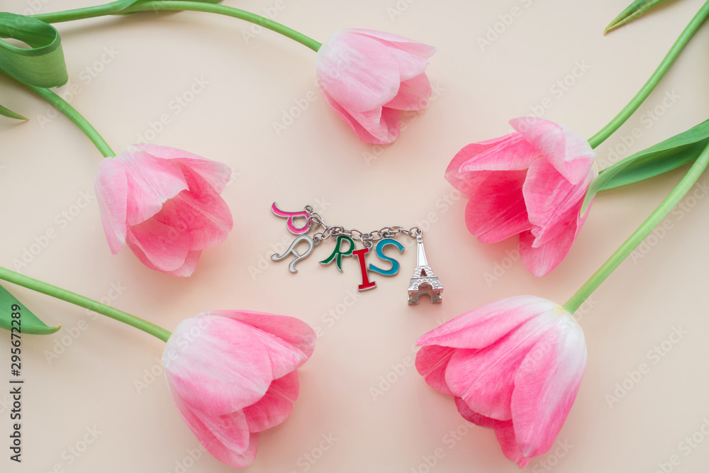 A close-up photograph of five pink flower buds around a keychain that looks like the Eiffel Tower with the inscription Paris. Top view on a beige background. A close-up photograph of five pink flower buds around a keychain that looks like the Eiffel Tower with the inscription Paris. Top view on a beige background.