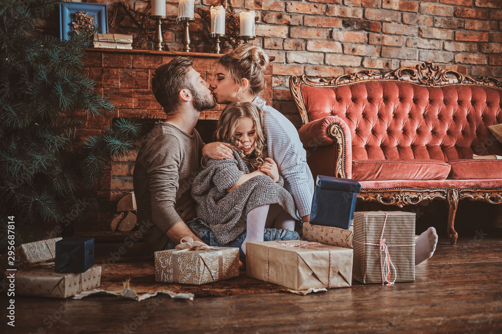 Surrounded by christmas gifts, mother and father are kissing, while holding their little daughter. - obrazy, fototapety, plakaty 