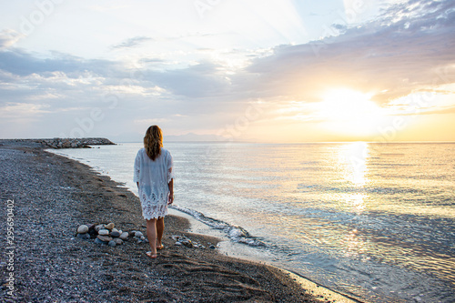A girl in white attire walks along the beach during sunrise.
