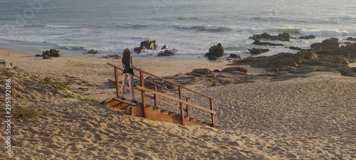 Escadas em madeira no meio da praia com uma rapariga a olhar para o horizonte