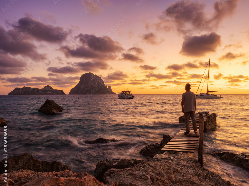 Fototapeta premium atardecer de otoño en la playa y embarcadero de Cala D´hort con vistas a Es Vedra. Hombre observando los barcos y el paisaje