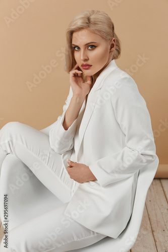 A fashionable young seductive woman with perfect body and with perfect blond hair in an elegant white suit posing in studio. Beautiful blonde model girl with full lips and trendy makeup.