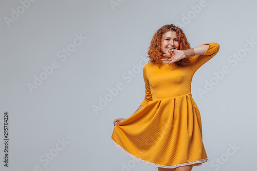 A red haired girl in a yellow dress on a light gray background smiles and touches her face