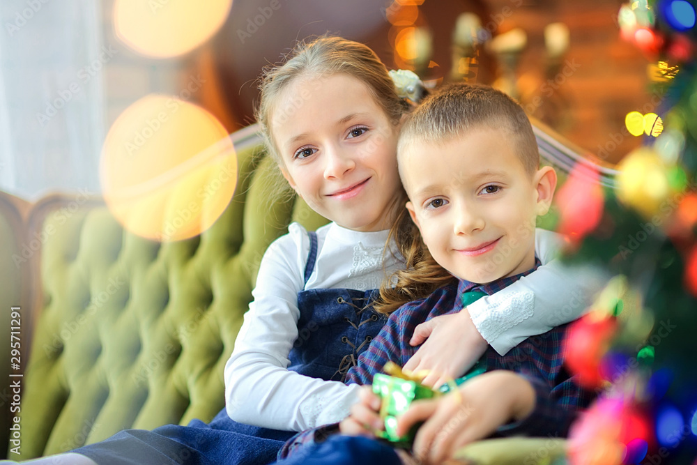 Christmas time, sister hugs little brother sitting on the sofa near the festive Christmas tree, smiling happy children Christmas time, sister hugs little brother sitting on the sofa near the festive Christmas tree, smiling happy children