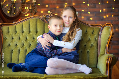 Christmas time, sister hugs little brother sitting on the sofa on the background of a garland, smiling happy children