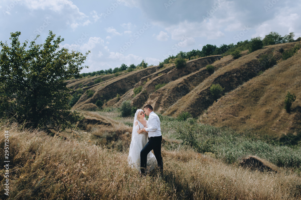 Wedding portrait of lovers newlyweds on a background of hills and sky with clouds. Stylish groom hugs a beautiful blonde bride. Photography and concept, emotions.
