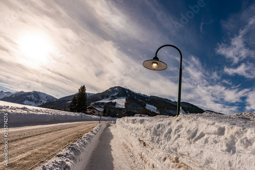 Lighting street lamp during the day on a snowy, empty street in winter. Waste of energy, unfriendly to the environment. Sun, mountains at background. Rohrmoos Untertal , Schladming Dachstein, Austria photo