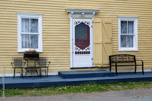 The exterior of an old historic building with yellow wooden clapboard, white screen door and white wooden glass pane windows. There are benches, chairs and a table on the deck of the house. 