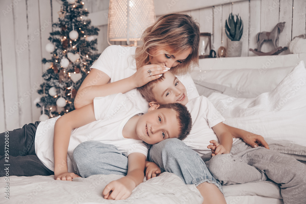 Happy loving family. Beautiful young mother and her two kids child boys playing, laughing and hugging in white Scandinavian style bedroom against the background of the Christmas tree