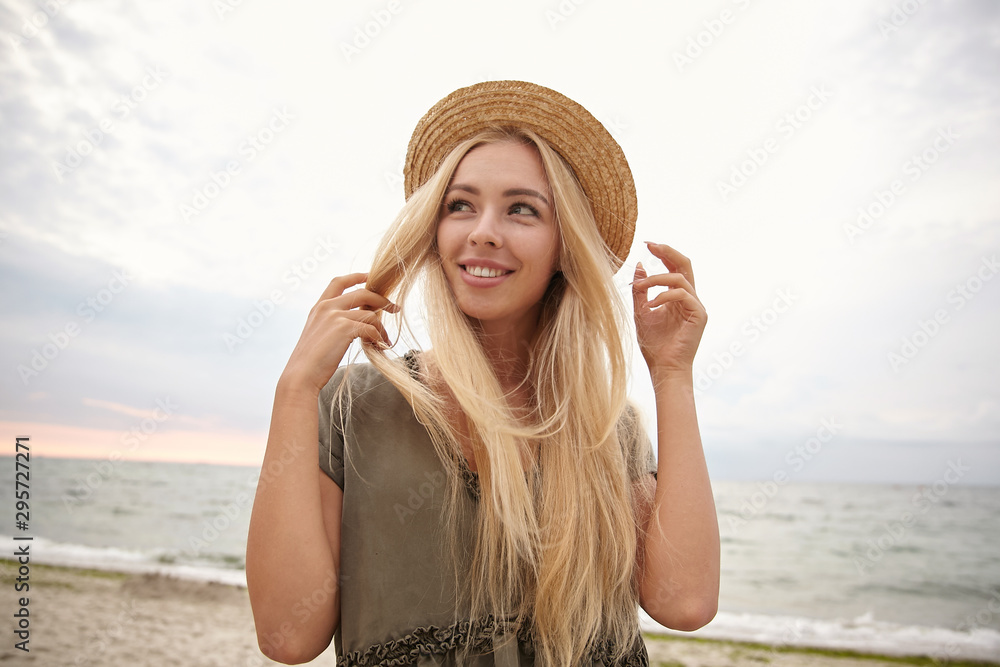 Portrait of attractive young blonde woman with long hair looking aside with charming smile, posing over seaside on overcast in casual dress and boater hat Portrait of attractive young blonde woman with long hair looking aside with charming smile, posing over seaside on overcast in casual dress and boater hat