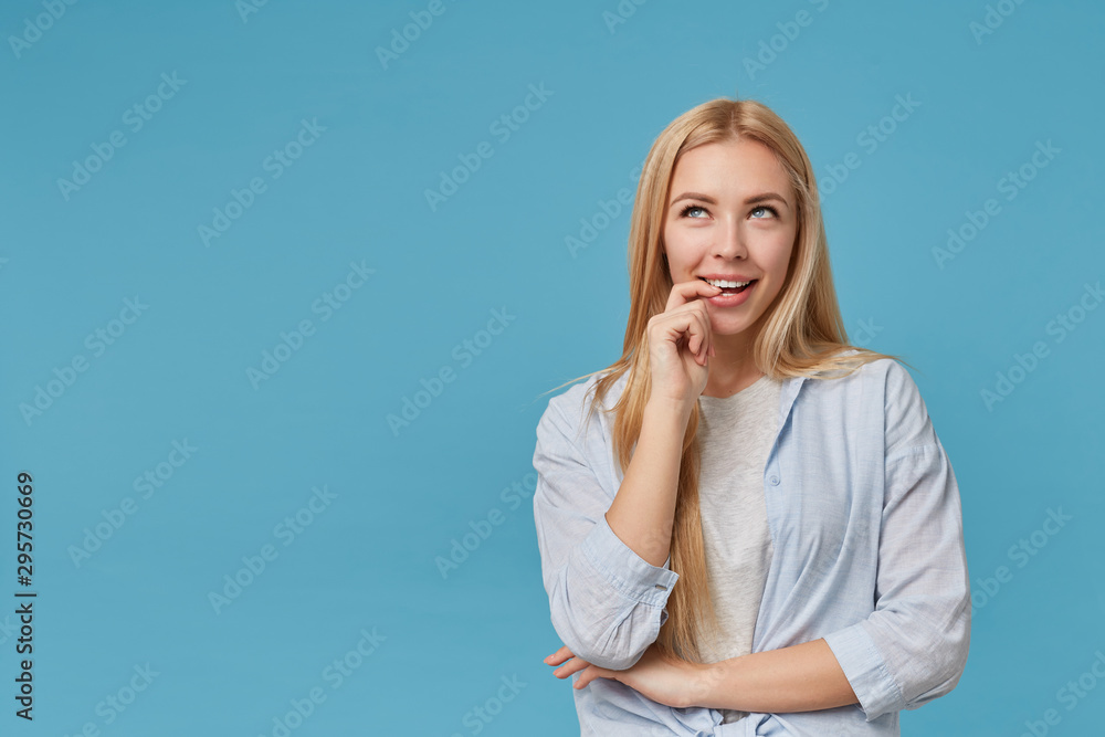 Joyful blonde long haired female wearing casual clothes, looking upwards dreamily and biting forefinger, showing her pleasant emotions, isolated over blue background Joyful blonde long haired female wearing casual clothes, looking upwards dreamily and biting forefinger, showing her pleasant emotions, isolated over blue background