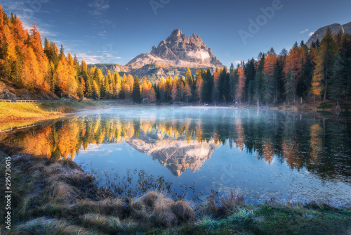 Lake with reflection of mountains at sunrise in autumn in Dolomites, Italy. Landscape with Antorno lake, blue fog over the water, trees with orange leaves and high rocks in fall. Colorful forest