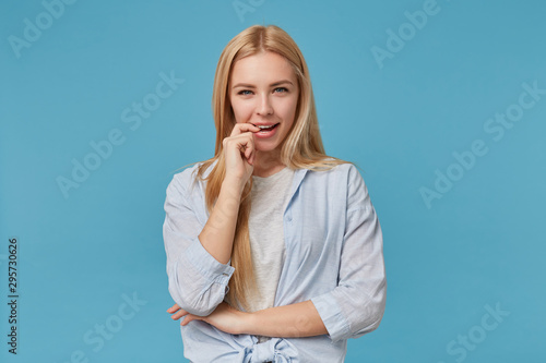 Portrait of flirty young long haired lady with casual hairstyle standing blue background, wearing blue shirt and grey t-shirt, looking at camera with squint and biting forefinger