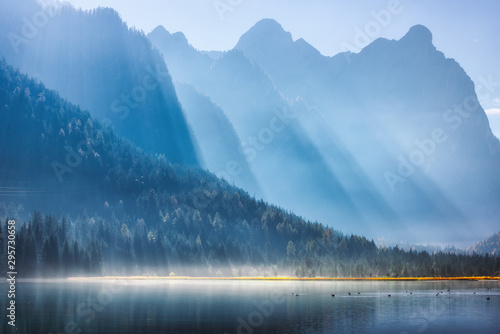 Fototapeta Naklejka Na Ścianę i Meble -  Majestic mountains in bright sunbeams and foggy lake at sunny morning in autumn.  Fall in Dolomites, Italy. Landscape with fog over water, high rocks and hills, forest and sunlight at sunrise. Nature