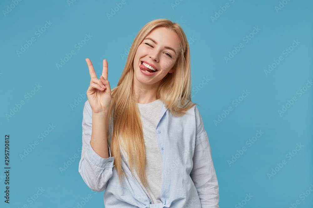 Joyful young blonde woman with long hair fooling over blue background in casual clothes, showing tongue cheerfully to camera and raising two fingers with victory gesture Joyful young blonde woman with long hair fooling over blue background in casual clothes, showing tongue cheerfully to camera and raising two fingers with victory gesture