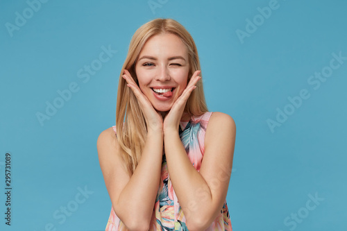 Funny indoor photo of young blonde lady winking at camera and showing tongue cheerfully, standing over blue background in romantic dress, keeping folded palms under her chin