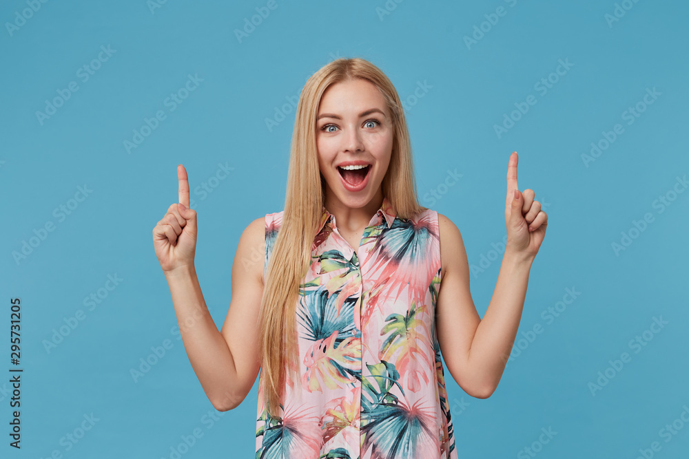 Happy blonde long haired female with casual hairstyle wearing romantic dress, showing upwards with forefingers while posing over blue background, smiling widely to camera Happy blonde long haired female with casual hairstyle wearing romantic dress, showing upwards with forefingers while posing over blue background, smiling widely to camera