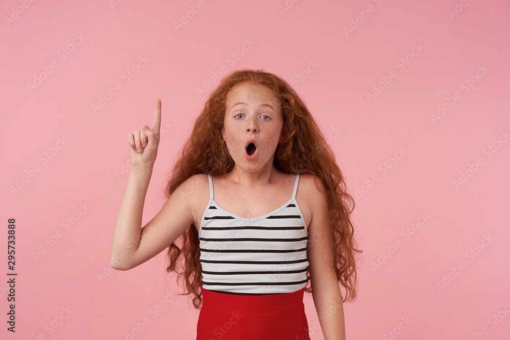 Studio photo of surprised curly redhead female kid with casual hairstle posing over pink background in elegant clothes, looking camera with wide mouth opened and pointing upwards with forefinger Studio photo of surprised curly redhead female kid with casual hairstle posing over pink background in elegant clothes, looking camera with wide mouth opened and pointing upwards with forefinger
