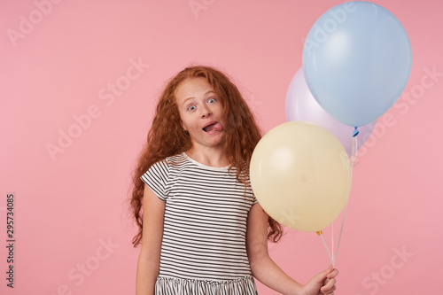 Funny portrait of redhead curly female kid in striped dress standing over pink background with air balloons, celebrating holday, showing tongue and making faces