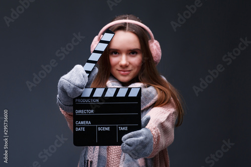 Young beautiful smiling girl in knitting pullover holding director's clapper board for making Christmas video in studio.Movie production clapper board.Lights, camera, action. Start of winter holidays. © YURII MASLAK