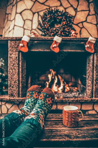 Stopy w wełnianych skarpetkach przy świątecznym kominku. Kobieta relaksuje się przy ciepłym ogniu z filiżanką gorącego napoju i rozgrzewa stopy w wełnianych skarpetach. Zamknij się na nogach. Zimowe i świąteczne wakacje koncepcji.