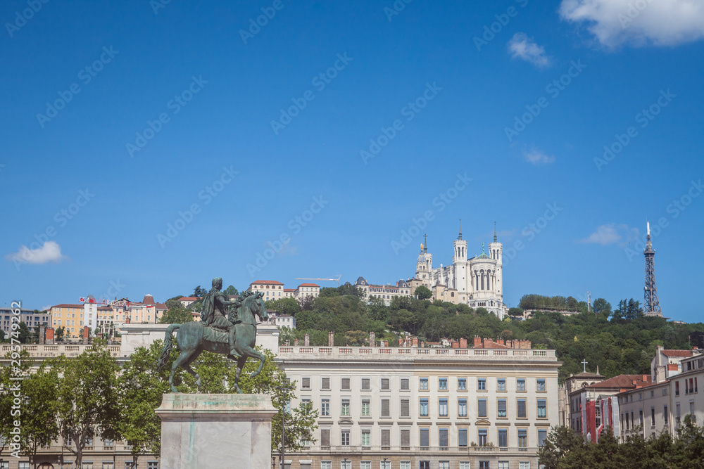 Roi Louis XIV statue on display on the Place Bellecour Square, in downtown Lyon, France, with the Basilique Notre Dame de Fourviere Church in background at sunset in summer