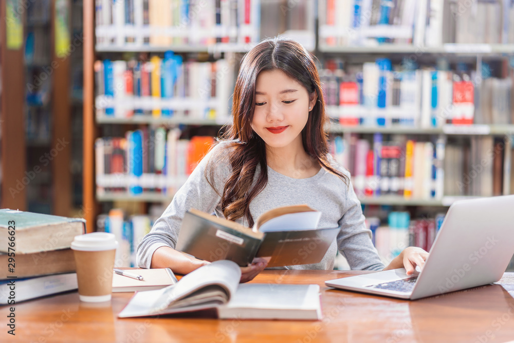 Asian young Student in casual suit doing homework and using technology laptop in library of university or colleage with various book and stationary over the book shelf background, Back to school