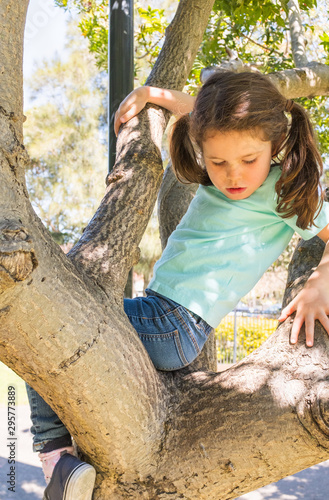 Vertical view of little girl in with hair in pigtails and blue t-shirt and jeans climbing tree in playground (selective focus)