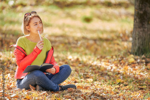 young woman sitting with a book on fallen leaves in the autumn park