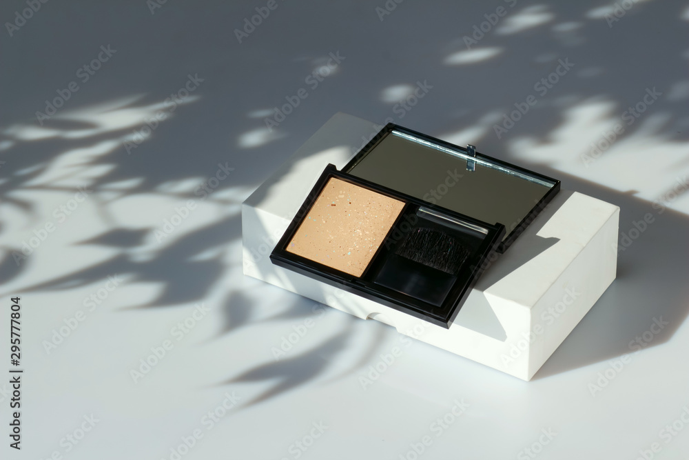 Fototapeta premium Face powder in a black case. Fashion cosmetics on a white background with a shadow of leaves. Open eyeshadow, compact foundation. Concealer face powder, blush. Cosmetics with natural ingredients.