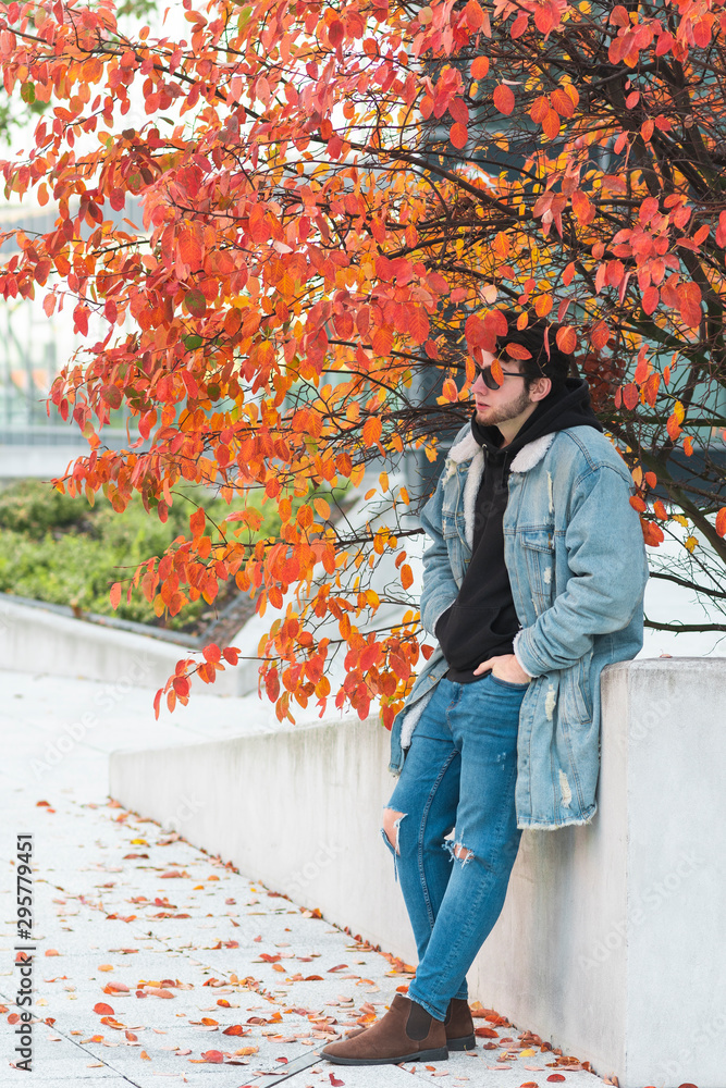 Obraz premium Young man standing in a concrete park under the tree, autumn fashion concept.