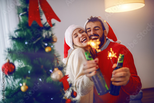 Attractive caucasian couple in love holding sparklers and beer bottles while standing in living room. Both having santa hats on heads. In foreground is christmas tree.