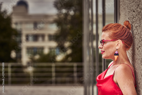 Portrait of a red-haired middle-aged woman in a red blouse standing against the wall. Cloudy summer day. Close-up.