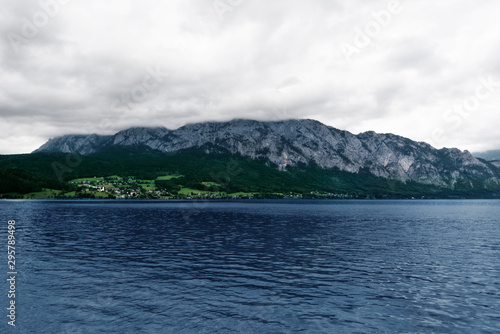 A village on other size of Kammersee lake. A cloudy day at Attersee lake at Salzkammergut area in Austria. There are a lot of green trees in the Alps mountains. The sky is full of grey clouds.