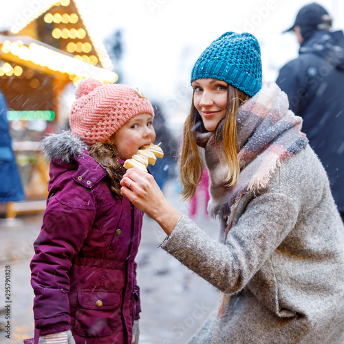 Young mother and daughter eating white chocolate covered fruits on skewer on traditional German Christmas market. Happy girl and woman on traditional family market in Germany, Munich during snowy day.