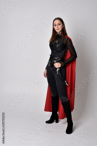 full length portrait of a pretty brunette woman wearing black leather fantasy costume with long red superhero cape. standing pose on a studio background.