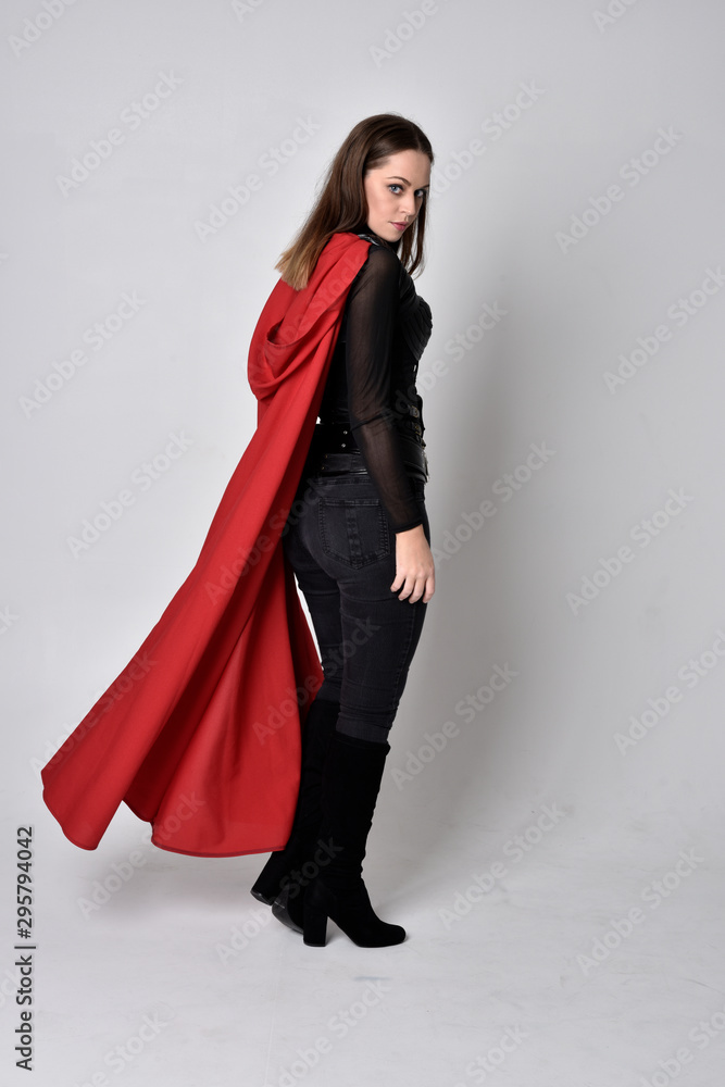 full length portrait of a pretty brunette woman wearing black leather fantasy costume with long red superhero cape. standing pose on a studio background. full length portrait of a pretty brunette woman wearing black leather fantasy costume with long red superhero cape. standing pose on a studio background.