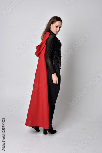 full length portrait of a pretty brunette woman wearing black leather fantasy costume with long red superhero cape. standing pose on a studio background.