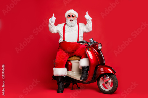 Portrait of his he nice bearded cheerful cheery funky confident Santa Claus hipster sitting on motor bike showing thumbup isolated on bright vivid shine vibrant red color background