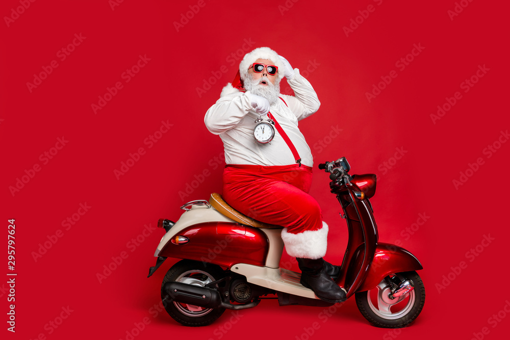 Profile side view of nice bearded worried funky Santa sitting on moped holding in hand clock countdown winter discount sale isolated on bright vivid shine vibrant red color background Profile side view of nice bearded worried funky Santa sitting on moped holding in hand clock countdown winter discount sale isolated on bright vivid shine vibrant red color background