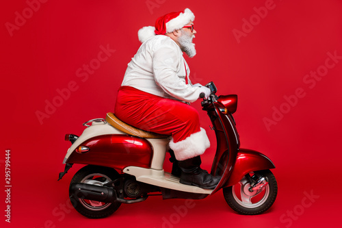Profile side view portrait of nice bearded serious confident Santa father riding motor bike delivering fairy tale dream shopping isolated on bright vivid shine vibrant red color background