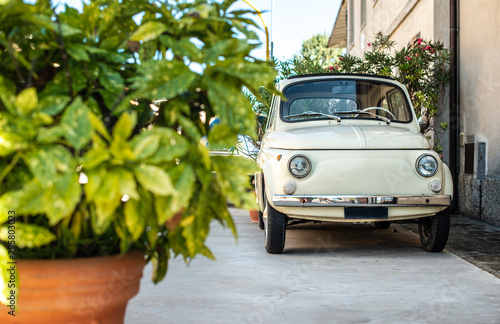 Small vintage italian car. Beige color old car in front of old house facade and flowers..