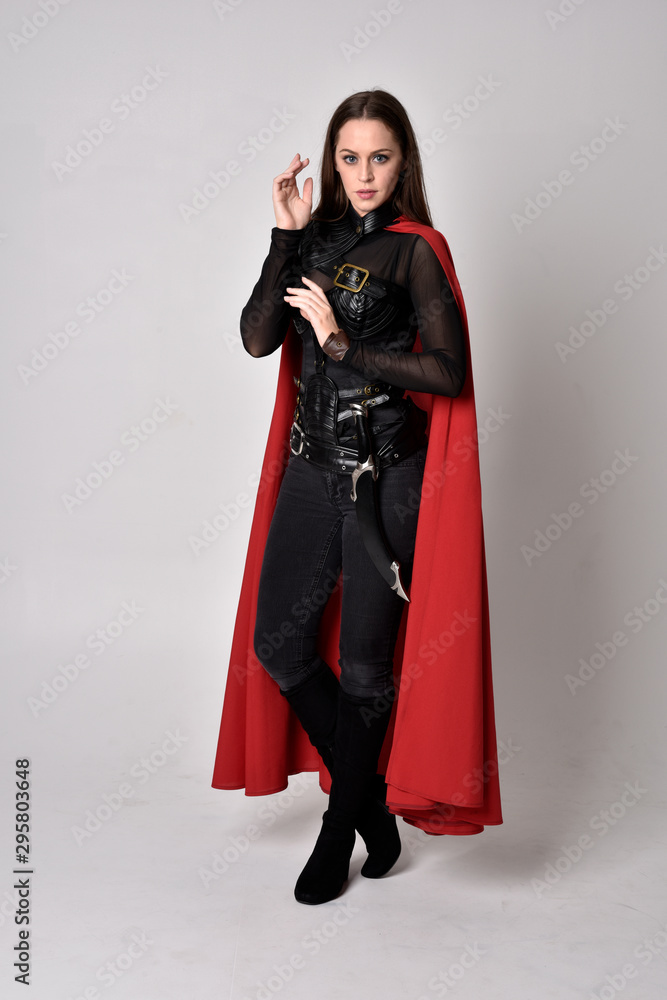 Naklejka premium full length portrait of a pretty brunette woman wearing black leather fantasy costume with long red superhero cape. standing pose on a studio background.