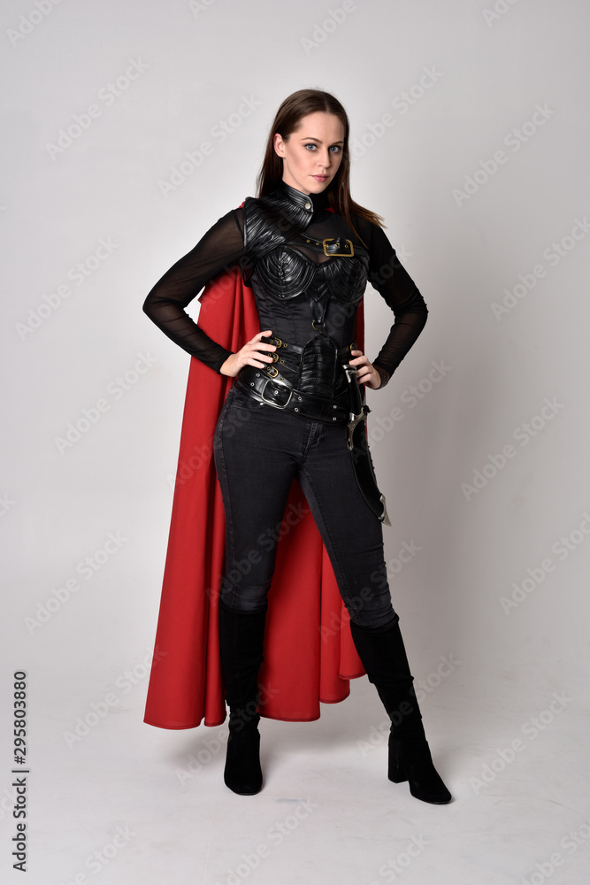 full length portrait of a pretty brunette woman wearing black leather fantasy costume with long red superhero cape. standing pose on a studio background. full length portrait of a pretty brunette woman wearing black leather fantasy costume with long red superhero cape. standing pose on a studio background.