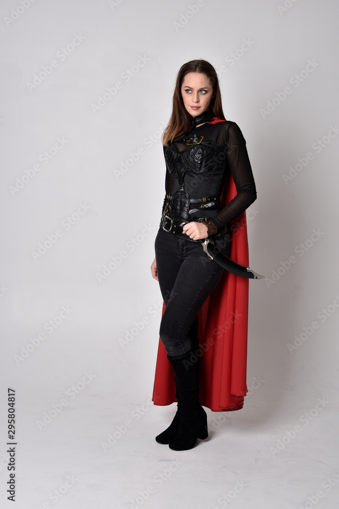 full length portrait of a pretty brunette woman wearing black leather fantasy costume with long red superhero cape. standing pose on a studio background. full length portrait of a pretty brunette woman wearing black leather fantasy costume with long red superhero cape. standing pose on a studio background.